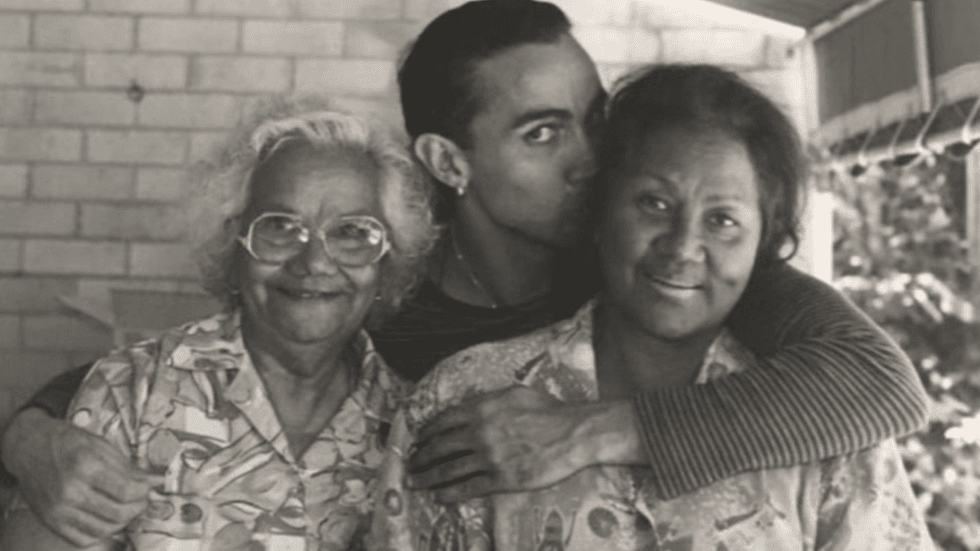 A black and white image of a young man standing behind his mother and grandmother