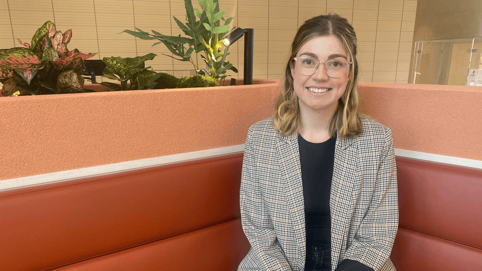 A blond woman with glasses in a blazer sits on a bench smiling at the camera