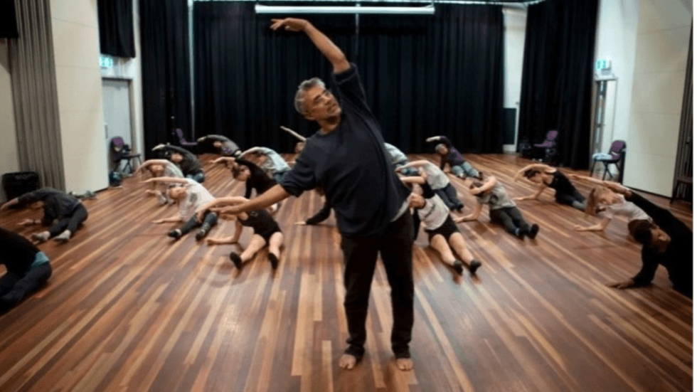 A choreographer in a studio with wooden floorboards surrounded by young dancers