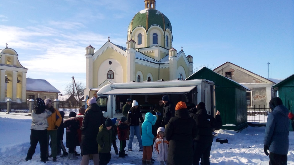 A crowd of people stand in the snow opposite a van, against a backdrop of a church