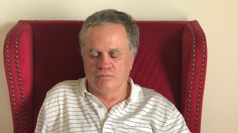 A grey-haired man sits on a red chair, wearing a white and black striped shirt.