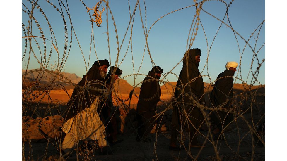 A group of Afghan elders walk outside a barbed wire barricade.