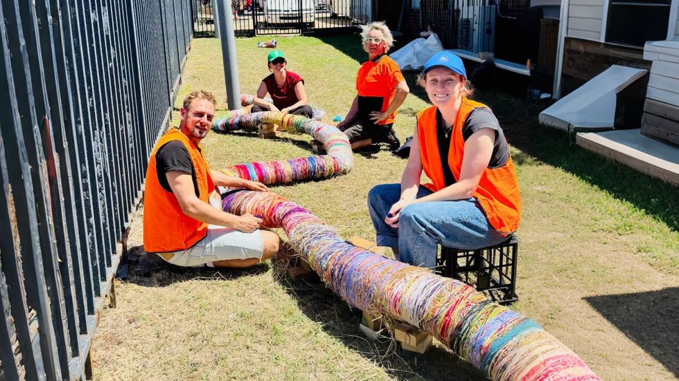 A group of people wearing orange vests weave an artwork