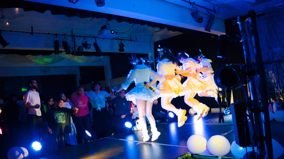 A group of young women dance on a stage facing a crowd in a darkented theater
