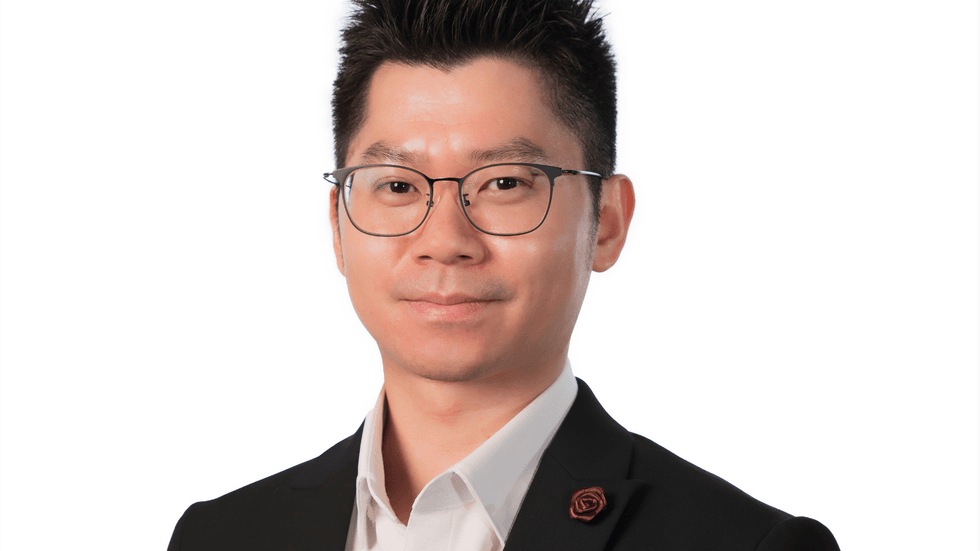 A man in a black suit with an open white shirt and black rimmed glasses