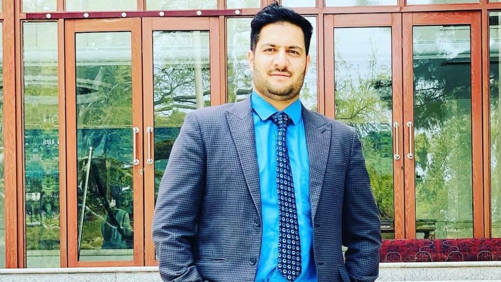 A man wearing a blue shirt and suit and tie smiles at the camera