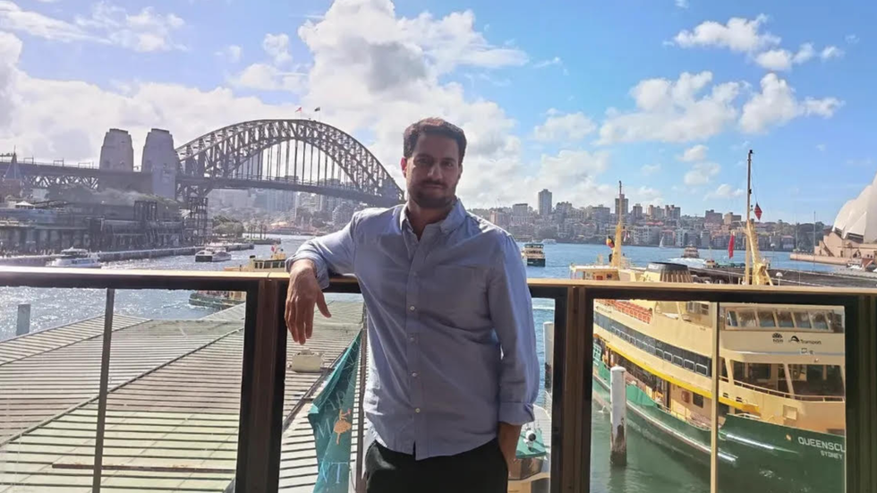 A man wearing a blue shirt stands at Circular Quay, his back towards Sydney Harbour bridge and the ferries