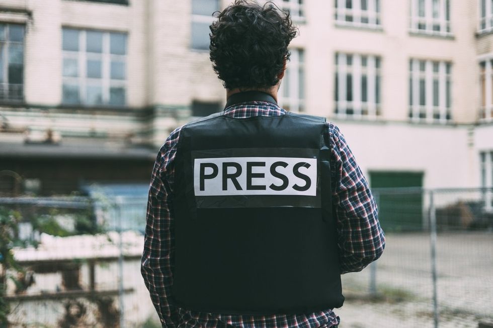 A man wearing a 'press' flak jacket faces away from the camera