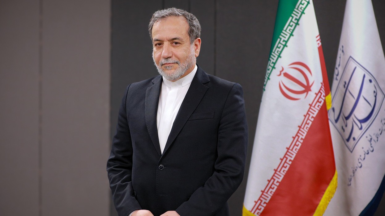 A man with a grey bear and wearing a suit in front of the Iranian flag