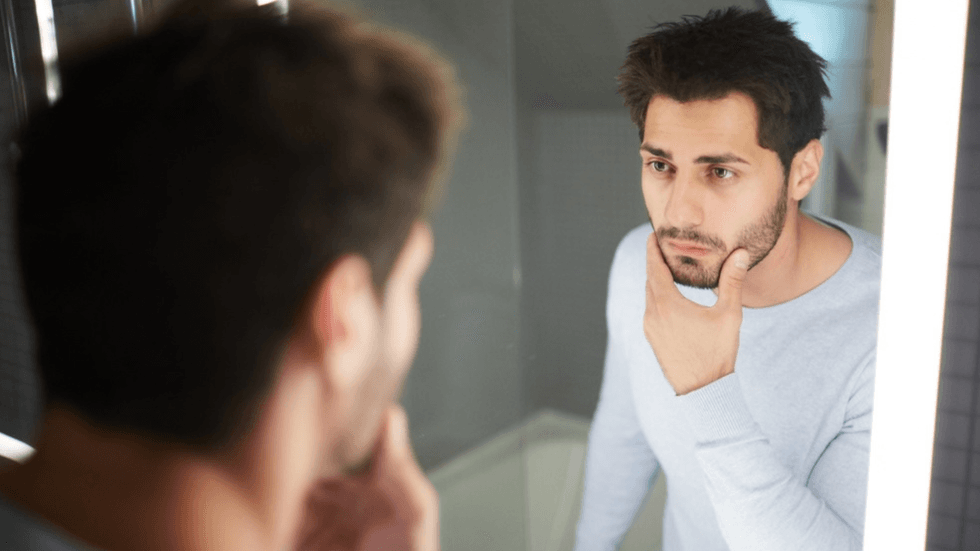 A man with dark hair and stubble stares into a mirror