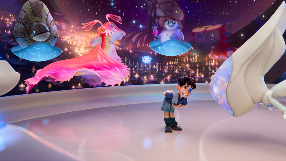 A screenshot from an animated film showing a small boy bowing in front of a humanoid figure. Behind him, three multicoloured aliens float
