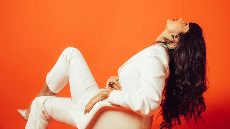 A side view of a woman with long dark hair sitting in a white chair. She's wearing a white suit and she has white high heels on.