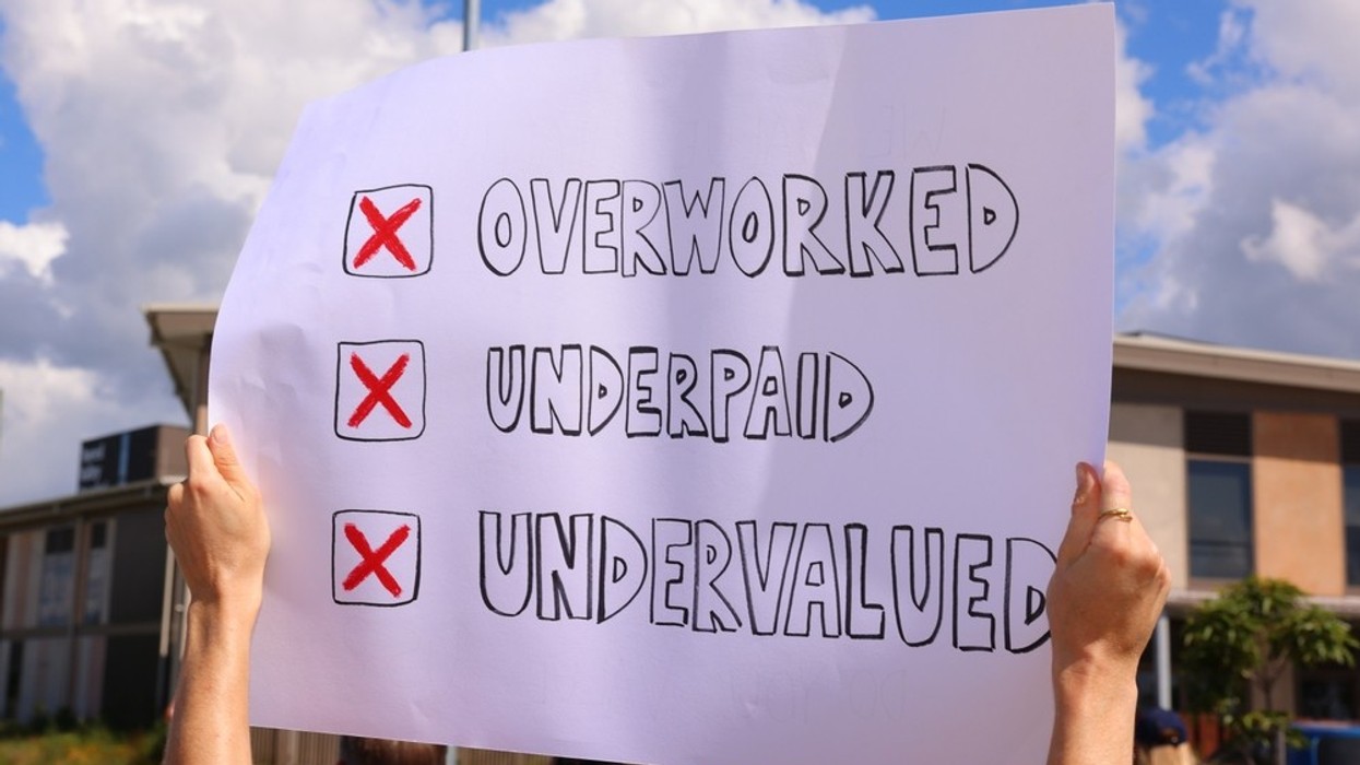 A sign held up by a person saying 'Overworked, Underpaid, Undervalued'.