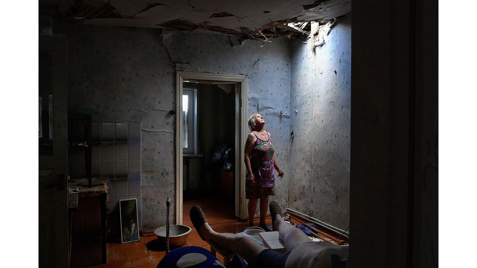 A Ukranian woman stares through the whole in her ceiling behind an ill man.