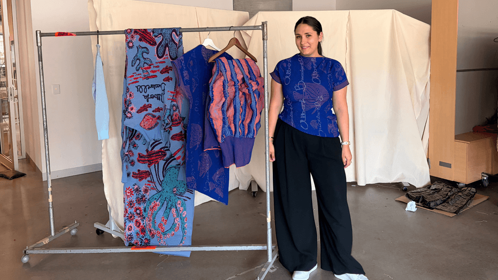 A woman in black pants and a bright blue top stands next to a clothes rack of knitted items.