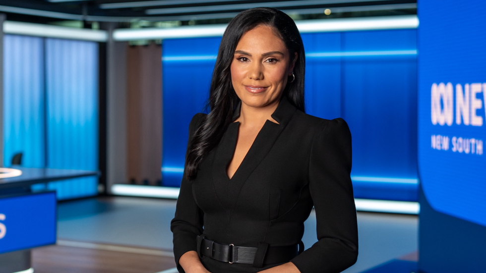 A woman smiles at the camera, against the backdrop of a tv studio, dressed in a black sheath dress.