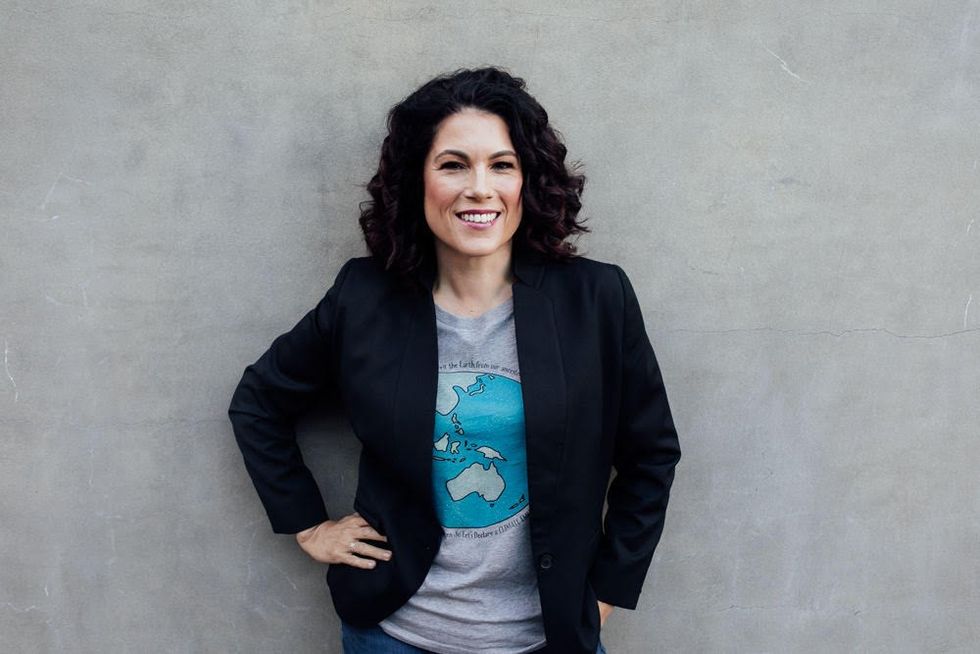 A woman with dark curly hair stands against a grey wall