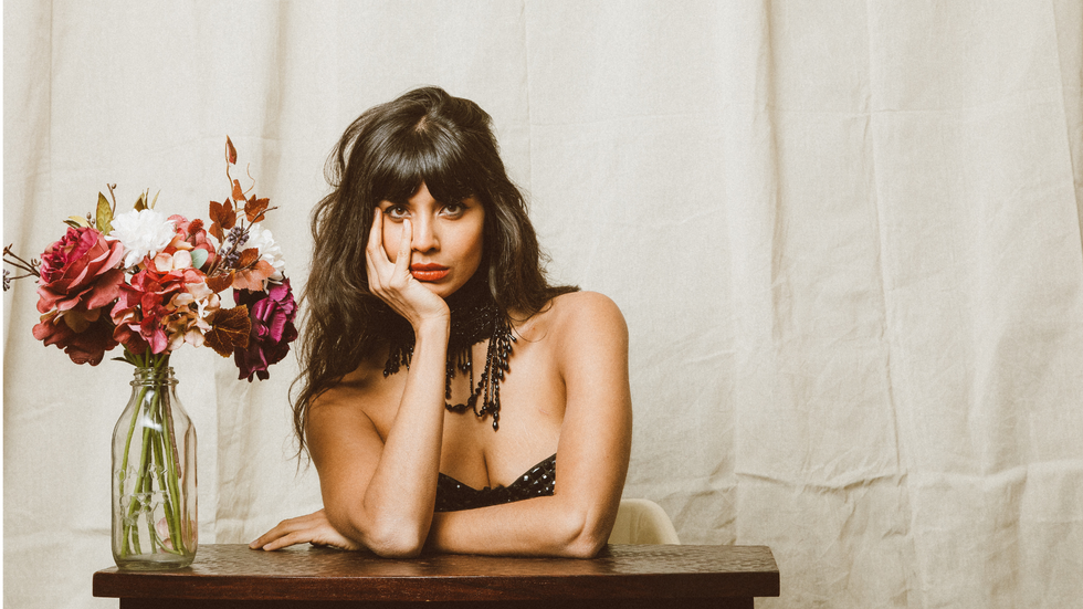 A woman with long dark hair wears a black bustier and a black necklace. She sits at a desk next to a vase of flowers