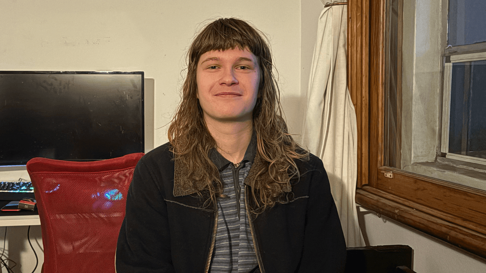 A young man with long hair sitting in a bedroom