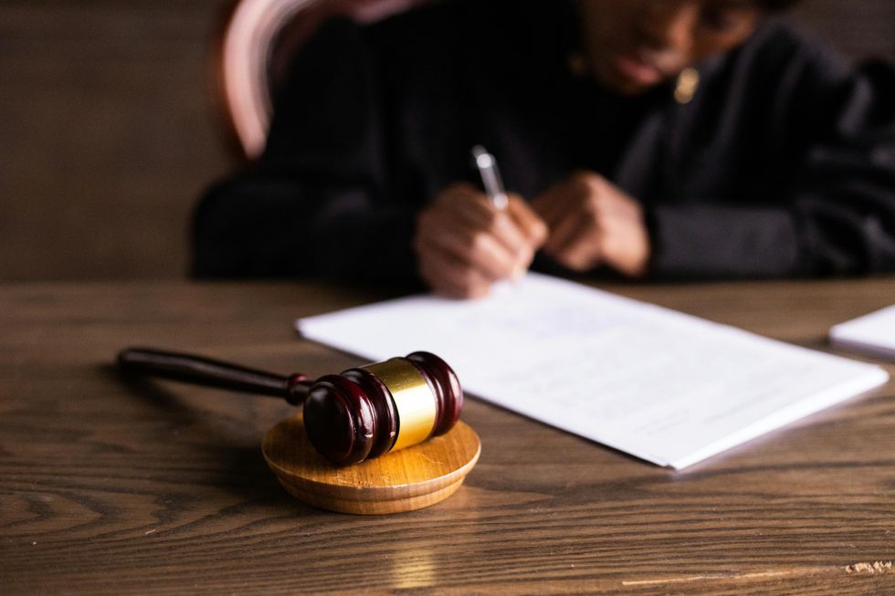 An image showing a gavel lying on a desk, while in the background a person fills out something on paper.