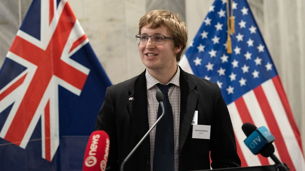 Head and shoulders image of a young man of Anglo appearance in a suit and tie at a podium with the Australian and US flags in the background.