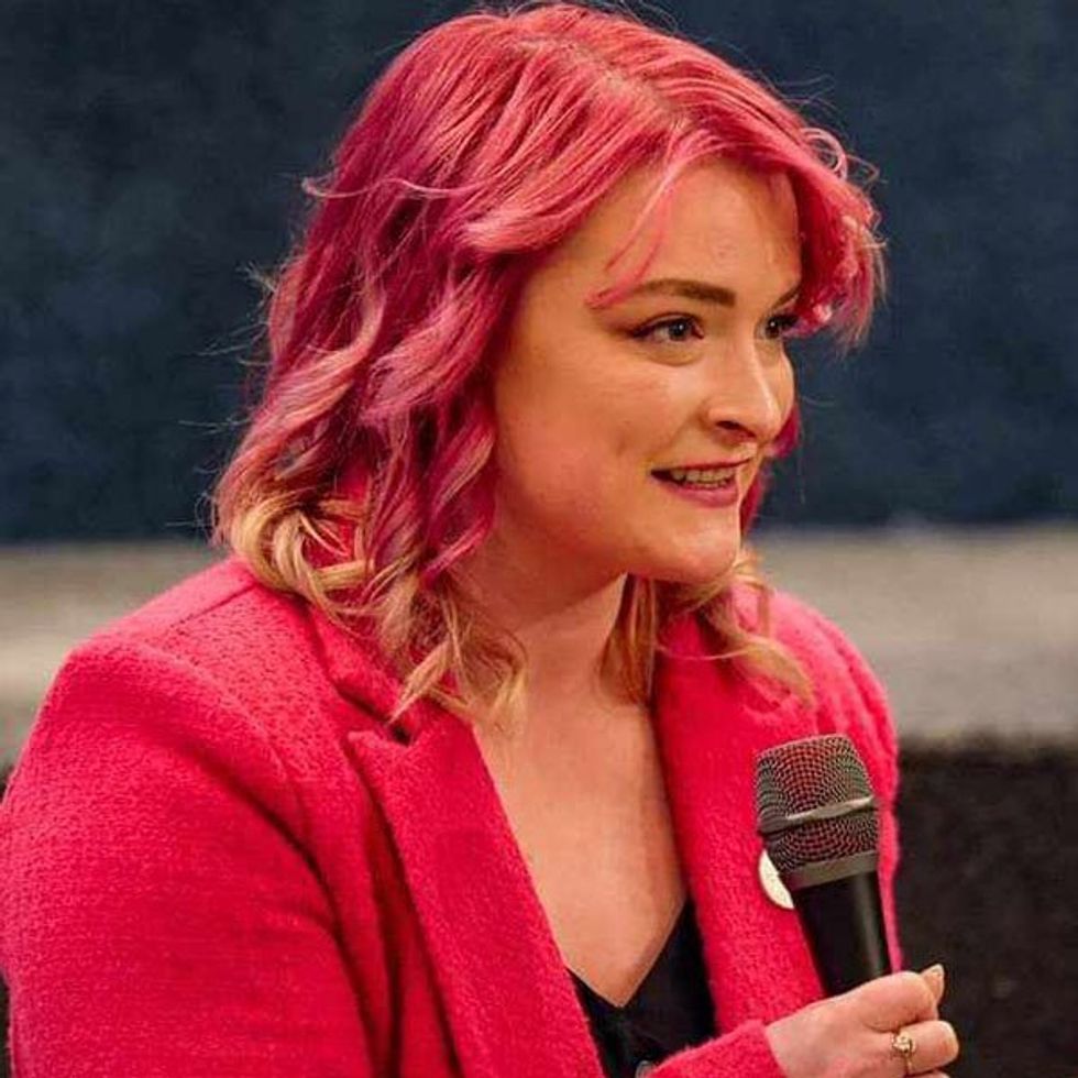 Head and shoulders image of a young woman with dyed red hair in a red jacket holding a microphone.