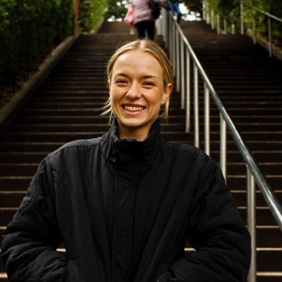 Image of a smiling young woman in a black jacket, from the waist up, with a set of steep stairs behind her.