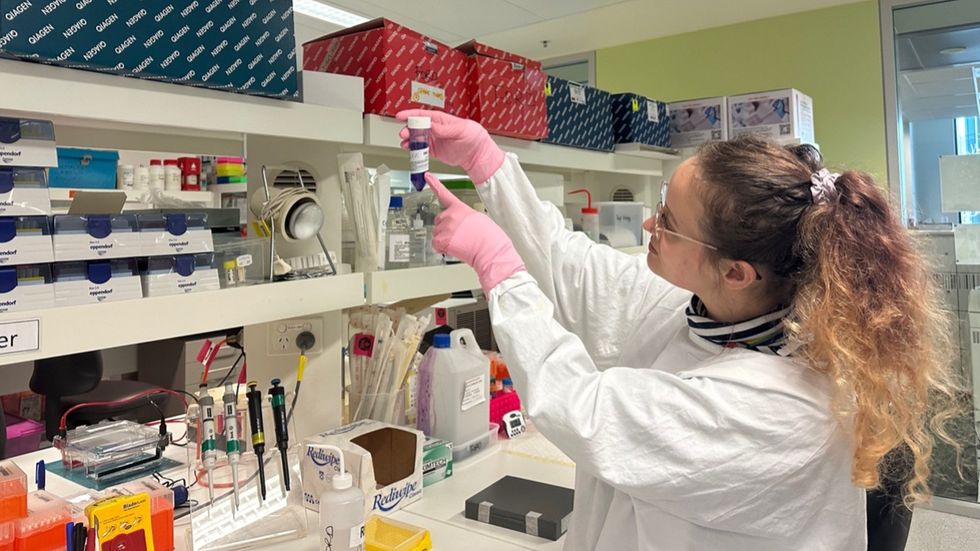 Image of a white-coated young female scientist in a lab pointing at a test tube.