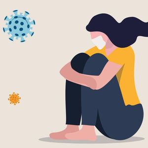 Illustration of a masked young woman sitting down with her arms wrapped around her raised knees. She is surrounded by floating COVID viral symbols.