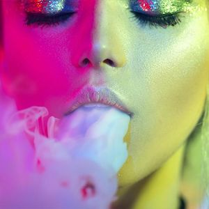 Image of a woman's face, in gilded make-up, blowing out smoke.
