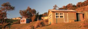 Image of three small fibro shacks on a hilllside bathed in the first soft light of day.