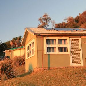 Image of three small fibro shacks on a hilllside bathed in the first soft light of day.
