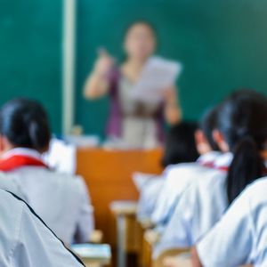 Image shot from behind of rows of students listening to a teacher at the front of the classroom.