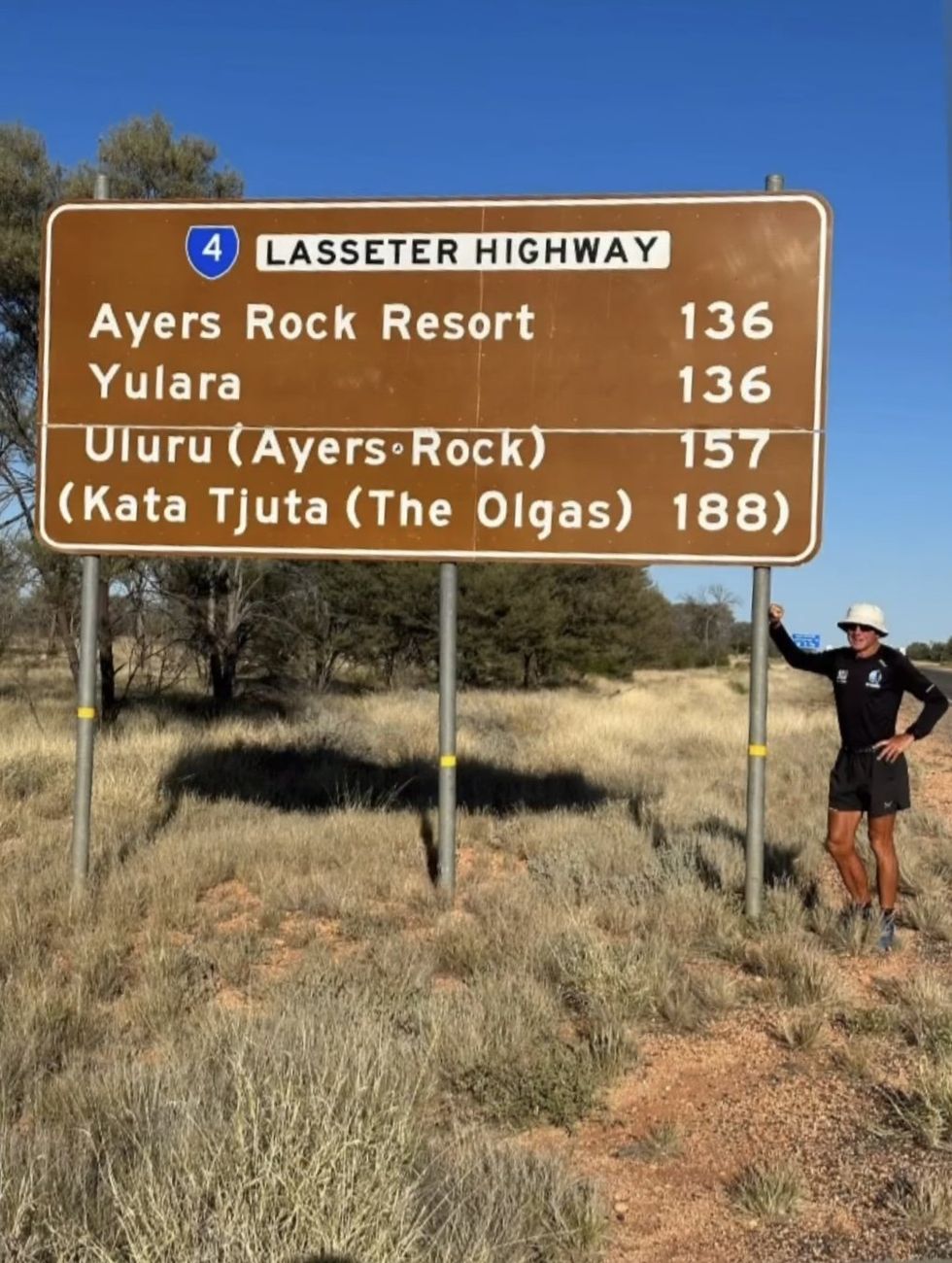 Pat Farmer standing next to the road sign to Uluru on the Lasseter Highway