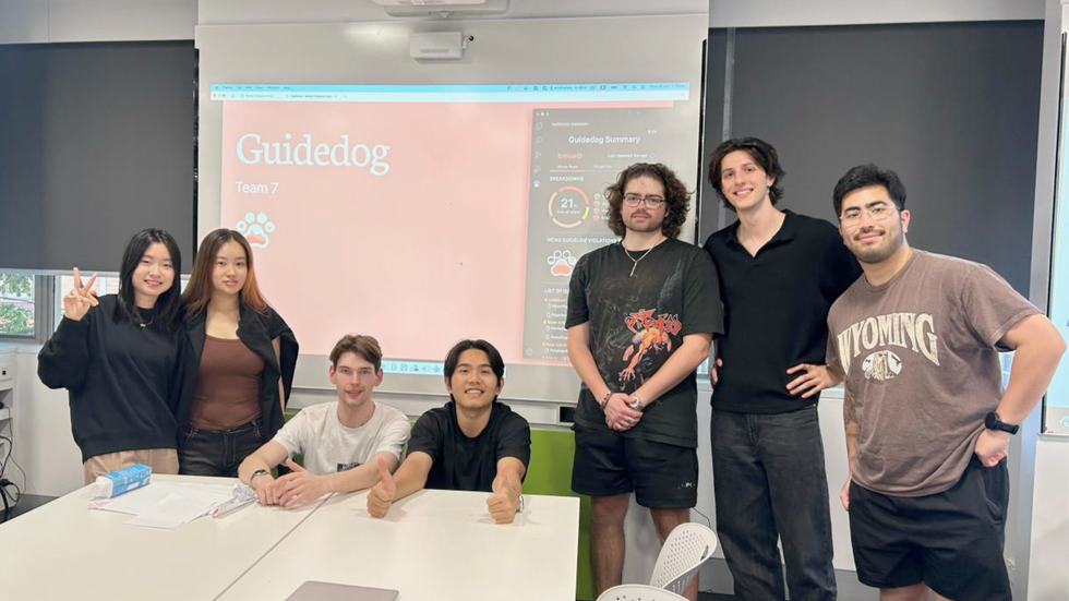 Seven university students smile in front of a projector screen that reads 'Guidedog'.