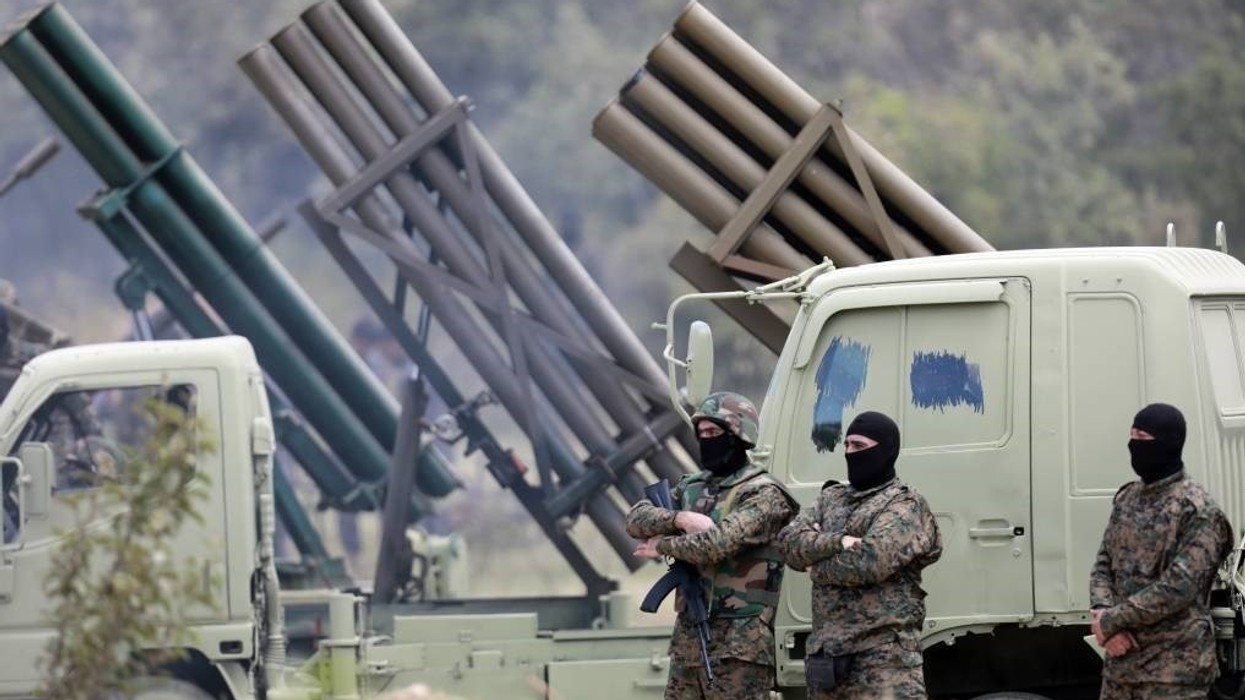 Three men wearing balaclavas and camouflage gear stand in front of rocket launchers.