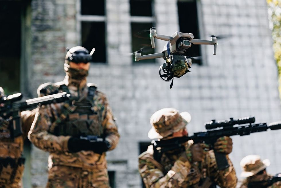 Two soldiers in camouflage holding guns and a drone in the air